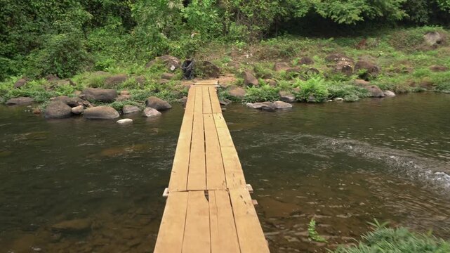 POV walking shot across a narrow wooden plank bridge over a rocky stream near Tad Champee waterfall on the Bolaven Plateau.