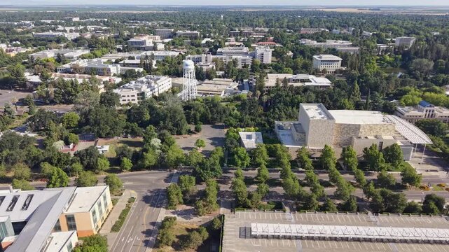 Expansive drone view of the University of California, Davis campus, showing modern academic buildings, lush green spaces, a distinctive water tower, and large parking structures on a sunny day