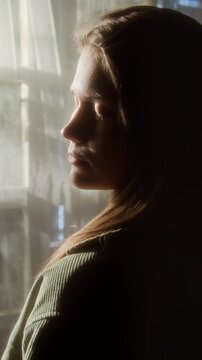 Vertical over shoulder shot of Caucasian young woman standing by window at home wind rustling hair and enjoying flow of fresh femininity accentuated by sunlight, story format