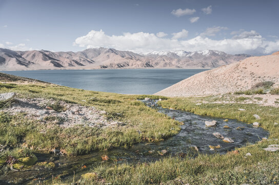 A mountain stream flows down a slope among green grass in sunny summer weather, flowing down to Lake Yashilkul, against the backdrop of a mountain range. Landscape background