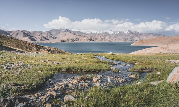 A mountain stream flows down a slope among green grass in sunny summer weather, flowing down to Lake Yashilkul, against the backdrop of a mountain range. Landscape background