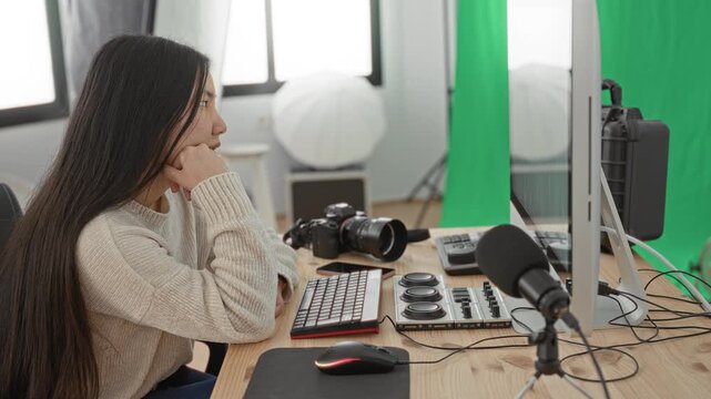 Woman at studio desk with camera and microphone, resting chin on fist while gazing at monitor and keyboard; contemplation creativity focus.