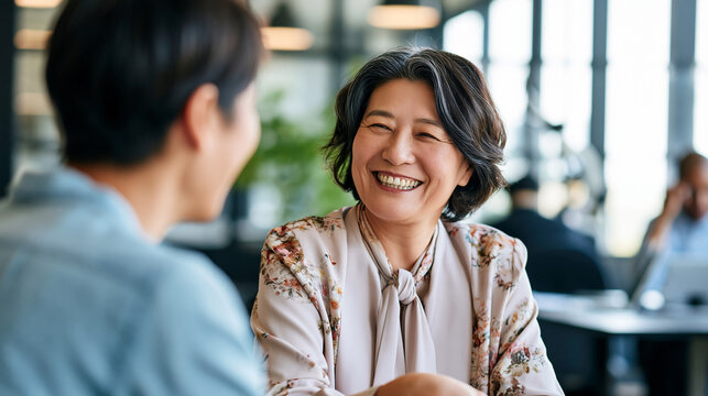A Japanese senior businesswoman smiles warmly while interviewing a calm young candidate in a bright modern office in soft daylight candidate interview open face modern office