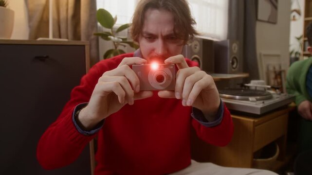Guy in red sweater, holding silver camera, intently framing shot against backdrop of record player during cozy home party
