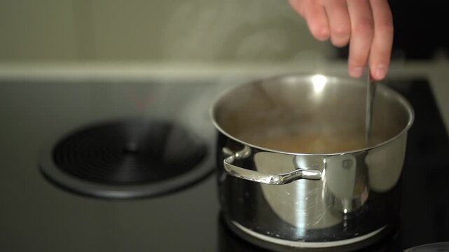 A metal pot sits on a black stove, pasta boiling inside while a man's hand stirs it, steam being drawn into the central extractor hood above. Cooking tutorial videos, kitchen appliance advertisements
