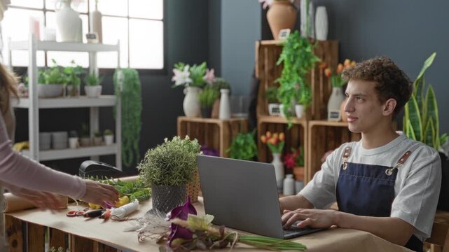 Man in apron types on laptop while woman leans forward with hand on table among flowers and vases in studio; warmth collaboration.