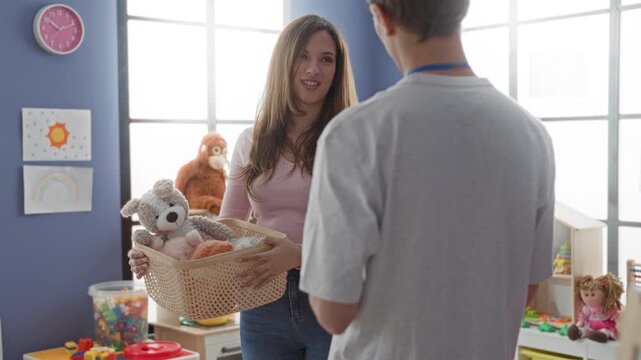 Woman holds basket of stuffed toys and smiles to man with clipboard while offering toys in a daycare building; care teamwork.