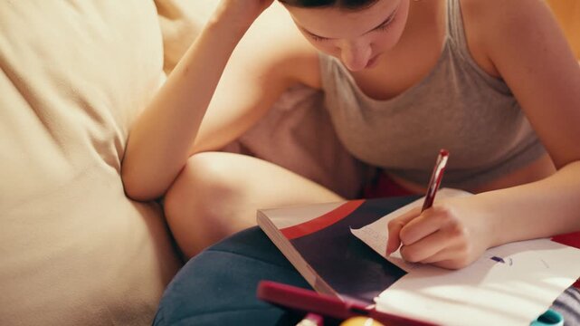 Deeply focused and worried high school girl making secret notes and tiny cheat sheets from her mobile phone, preparing for a test while sitting on a couch under warm indoor lighting.
