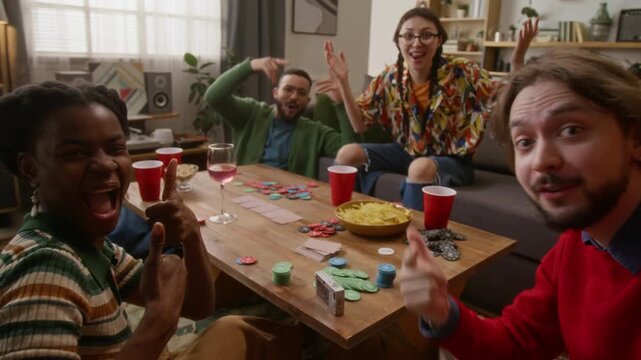 Portrait shot of young adult bearded man recording vlog of having fun with his friends, playing poker, drinking and eating snacks in cozy living room