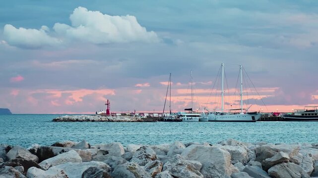 White shoreline rocks frame a yacht and red beacon near the Naples breakwater at dusk, under pastel clouds and calm Mediterranean water.