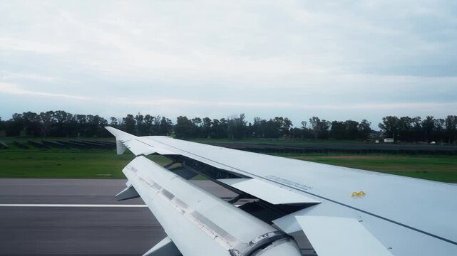 Passenger window view of a jet wing rolling on the runway after landing, with flap panels extended, green airfield beyond, and a calm overcast sky.