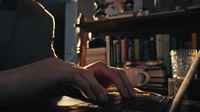 Man's hands typing on laptop keyboard in warm moody home interior at night.