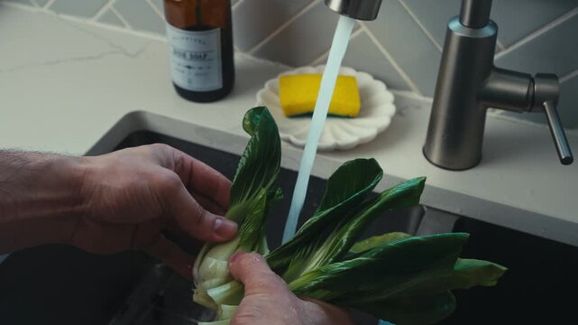 Man's hands rinsing fresh bok choy under tap water in modern black kitchen sink.