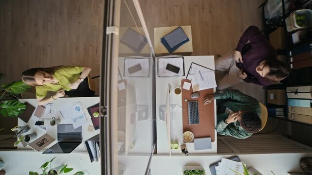Overhead shot of three diverse office employees sitting at their desks filled with paperwork and doing warmup exercises during routine workday in contemporary office environment