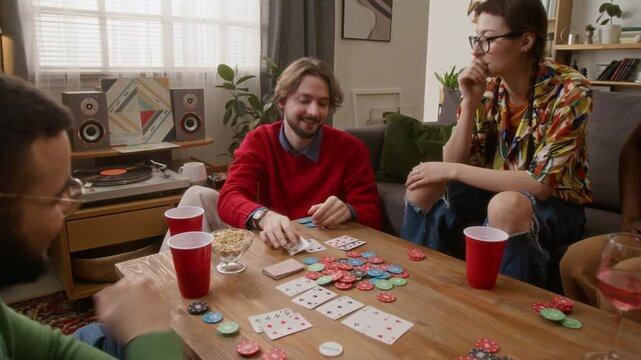 Young bearded man winning in poker and grabbing all playing chips, enjoying friendly gathering at home in cozy living room