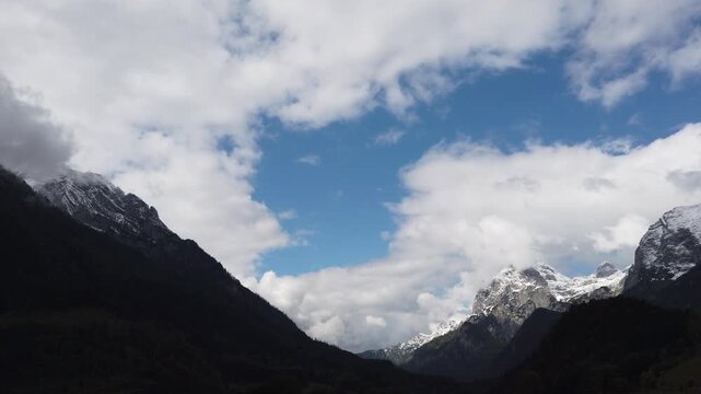 Snow-covered alpine peaks under a blue sky with clouds and dense forest landscape in the mountains