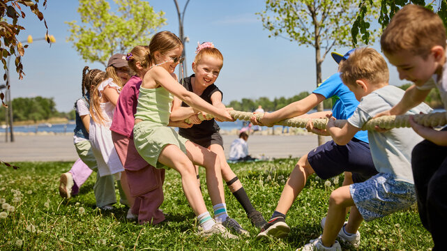 Children playing tug of war on grass during sunny outdoor summer game. Concept of outdoor teamwork, school camp, childhood leadership, group motivation, summer recreation and active lifestyle ads.