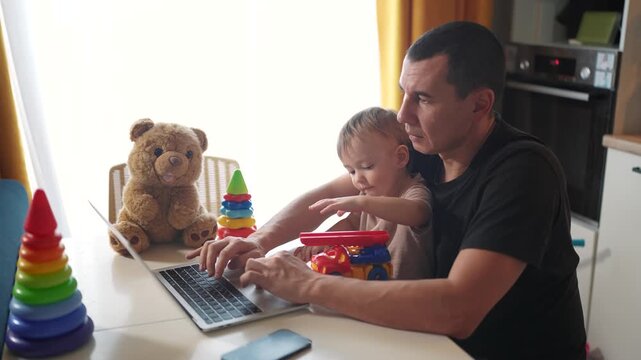 father working from home a remotely with baby son in his arms. pandemic remote work business concept. father tries to work at fun home in kitchen, baby children interfere sitting on their hands