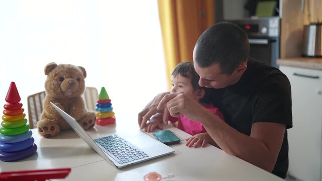 father working from home a remotely with baby daughter in his arms. pandemic remote work business concept. father fun tries to work at home in kitchen, baby children interfere sitting on their hands