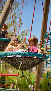 Girls giving high five on playground nest swing in sunny park. Concept of friendship, joyful childhood, outdoor play, school break, summer camp promo, family lifestyle and childcare content.
