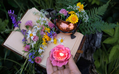 Obraz premium hand with burn floral shape candle, magic book, copper pot and wild flowers in forest, abstract nature background. Green witch, practical magic, esoteric ritual, witchcraft. close up