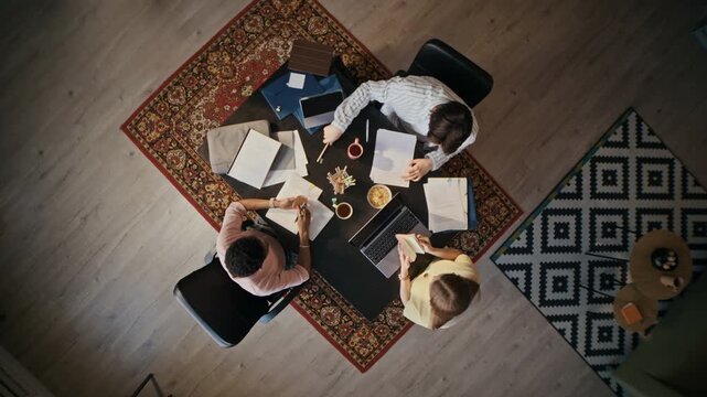 Overhead view of three young professionals working together with paperwork and digital devices during collaborative meeting in modern office