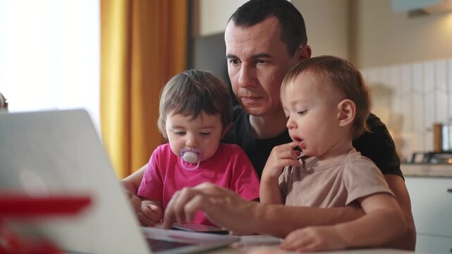 father working from home a remotely with two baby in his arms. pandemic remote work business concept. father tries to work at home in kitchen, baby children interfere sitting on their fun hands