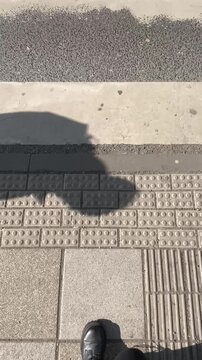 Pedestrian walking over tactile sidewalk tiles with guidance bumps and grooves, visible shoes and shadow crossing textured pavement, urban commuter. POV first person view