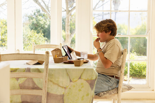 12 year old boy eating a box lunch in his kitchen while using tablet computer
