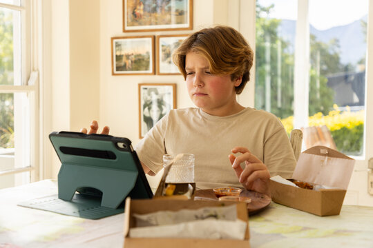 12 year old boy eating a box lunch in his kitchen while using tablet computer