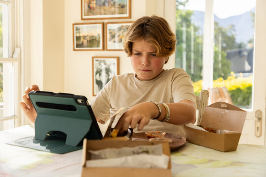 12 year old boy eating a box lunch in his kitchen while using tablet computer