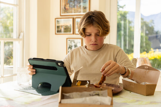 12 year old boy eating a box lunch in his kitchen while using tablet computer