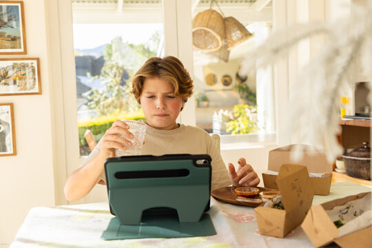 12 year old boy eating a box lunch in his kitchen while using tablet computer