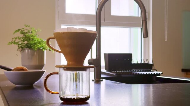 Fresh filter coffee slowly drips into a glass mug during a pour-over brewing process in a bright modern kitchen. Warm sunlight streams through the window while a ceramic dripper, paper filter
