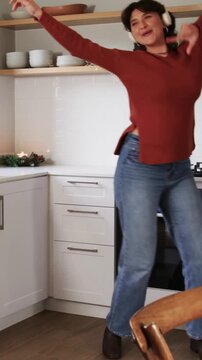 Vertical video: Dancing woman hearing music starting on headphones in kitchen, passing gas cooktop