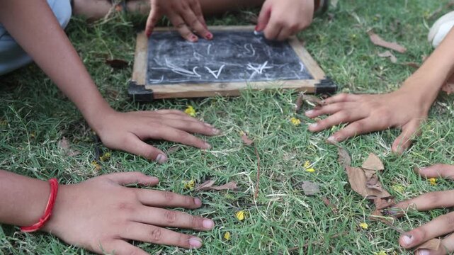 Indian village children sitting on green grass while playing and drawing together on a small blackboard during summer. Childhood friendship, outdoor activity, slum village poor india.