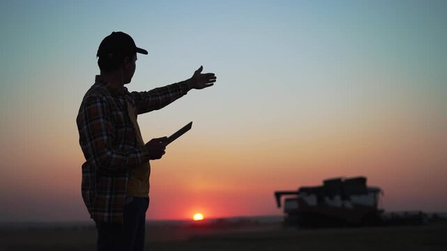 Agriculture. Harvester harvests wheat field at sunset. Silhouette of farmer with tablet in wheat field.Harvesting at sunset.Agricultural business.Farmer controls harvesting of wheat grain by combine
