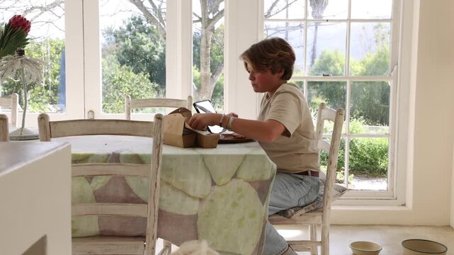 12 year old boy eating box lunch in kitchen using tablet computer