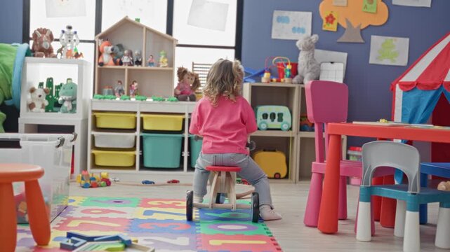 Girl kneels by a red striped play tent and reaches inside near a colorful children's table and chairs in a building playroom; playful curiosity.