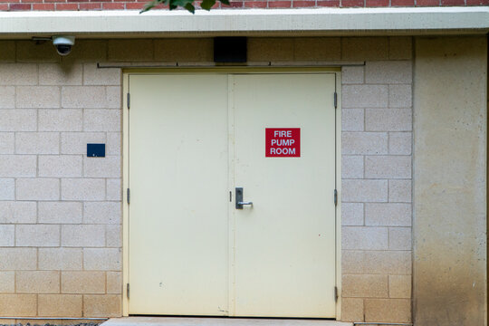Exterior view of a beige double door marked "FIRE PUMP ROOM" on a cinder block building, featuring a security camera for surveillance and emphasizing industrial safety.