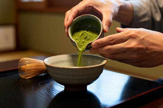 Person pouring matcha green tea powder into bowl on traditional wooden tray with bamboo whisk