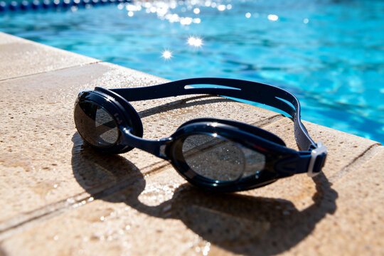 Swimming goggles resting on wet poolside surface with bright blue swimming pool in background