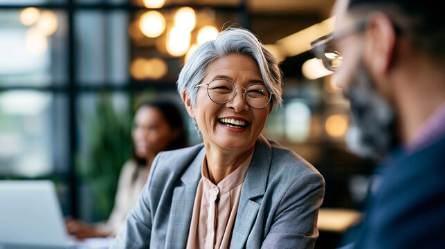 A mature Asian woman over fifty leads softly a bright meeting in a modern office wearing refined tailoring in natural daylight meeting lead open face office location natural