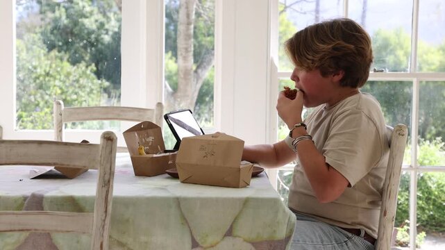 12 year old boy eating box lunch in kitchen using tablet computer
