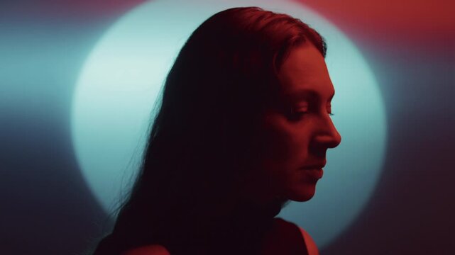 Pensive woman illuminated by red light looking down thoughtfully against blue luminous circular backdrop. Side profile view, zoom out shot