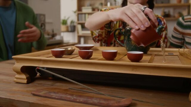 Low angle close up tilt up shot of young woman using gaiwan against backdrop of tea bowls, hosting tea ceremony for her friends