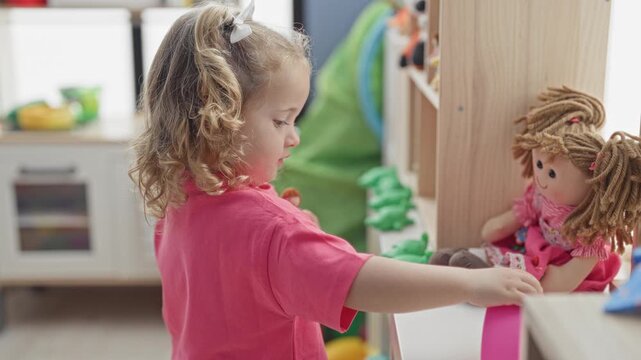 Little girl touching colorful abacus beads and holding a rag doll while reaching across a play table in a building; curiosity learning focus.
