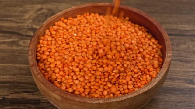 Pouring raw red lentils filling a rustic wooden bowl on a dark wood table