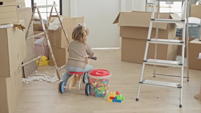 Little girl riding a wooden toy bike, holding handlebars and steering past stacked moving boxes in building; playful curiosity.