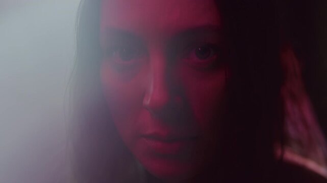 Close up portrait of young woman staring at camera under dramatic red light in dark studio with flickering flares
